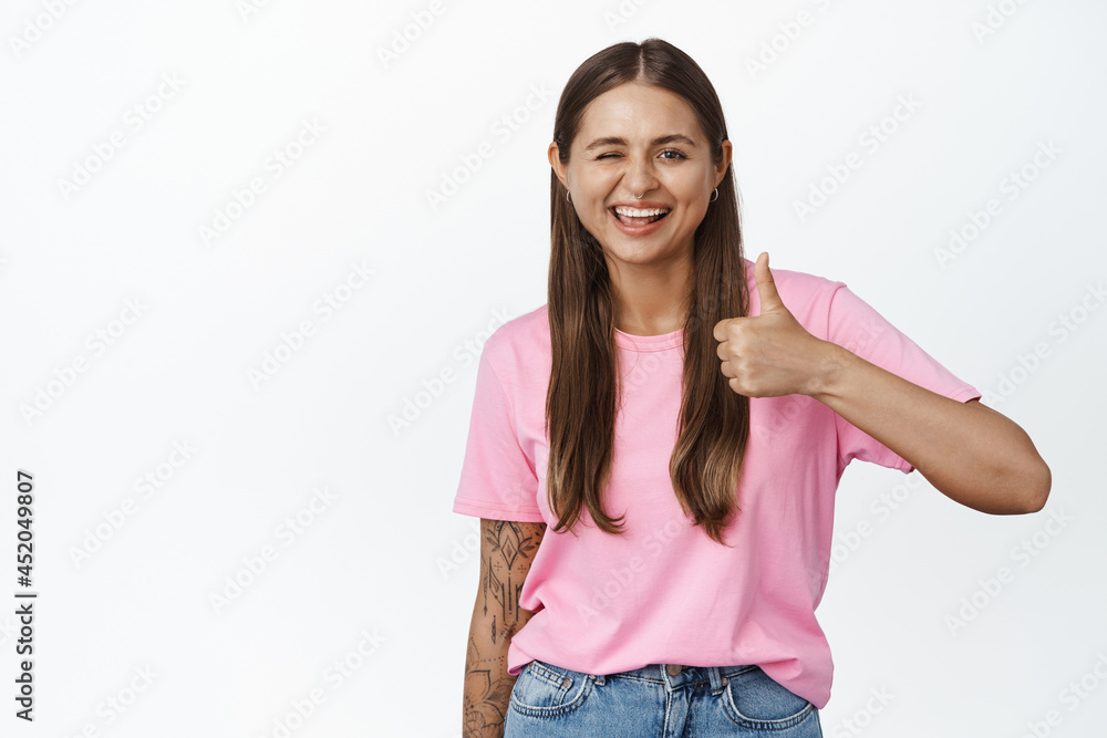 Winking girl smiling and showing thumbs up, wearing pink tshirt, standing over white background Winking girl smiling and showing thumbs up, wearing pink tshirt, standing over white background