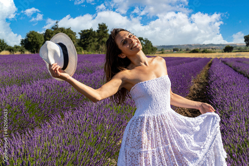 Portrait of a happy young woman playing with her hat in a blooming lavender field. Her white dress stands out against the purple color of lavender flowers.