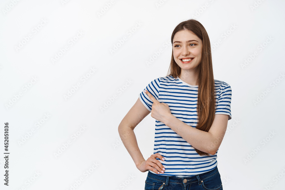 Happy and confident girl pointing, looking left with pleased smile, holding hand on waist, standing against white background Happy and confident girl pointing, looking left with pleased smile, holding hand on waist, standing against white background