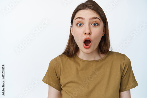 Face of surprised beautiful girl, saying wow, looking impressed at camera with oval-shaped mouth, standing in awe against white background