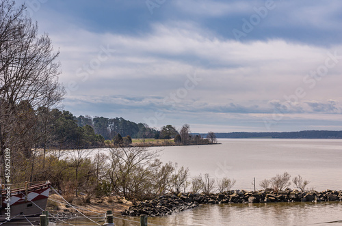 Jamestowne, VA, USA - April 1, 2013: Historic site. Gray water James River under blue cloudscape with brown and green foliage trees on shoreline. Bow of old boat at landing spot. photo