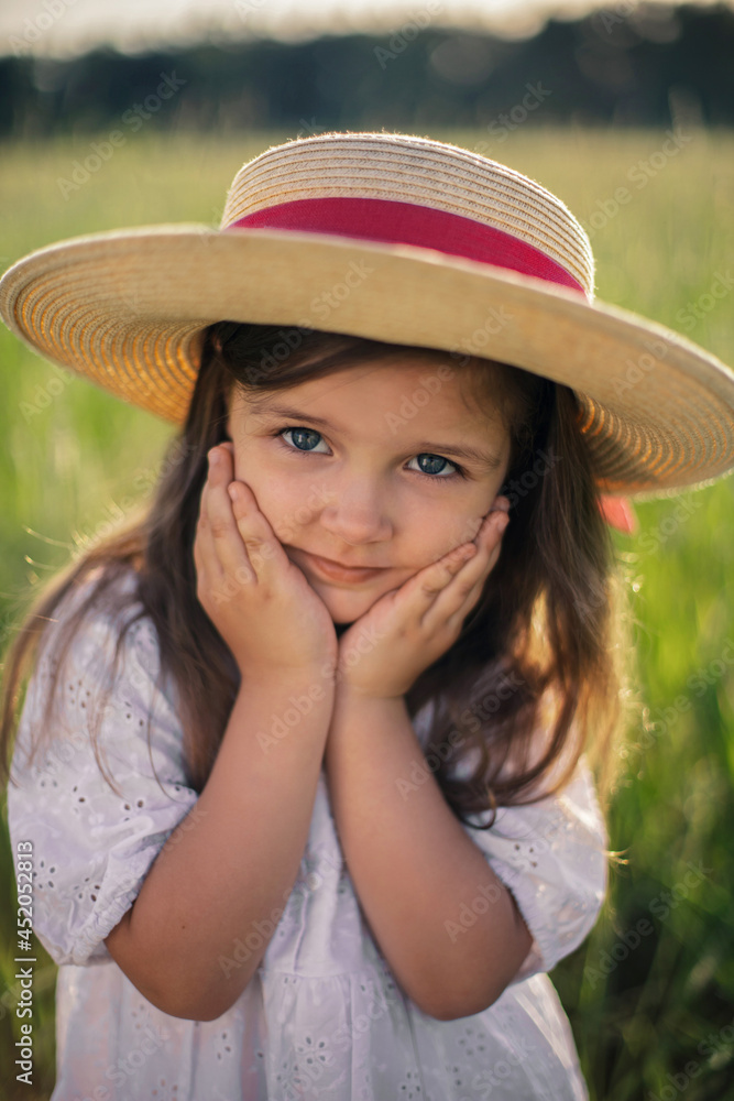 portrait of a beautiful girl in a straw hat standing in a field with green grass at sunset and looking the camera