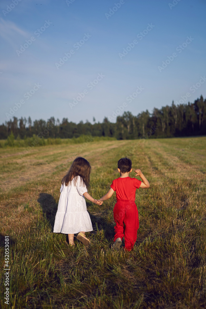 friend a boy in red clothes and a girl in a white dress walk through mown field at sunset in summer