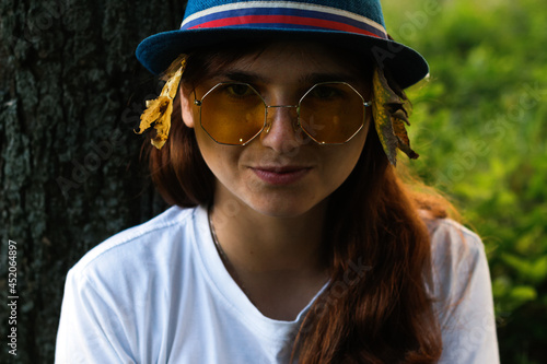 Defocus close-up portrait of a funny dreamy young woman with brown hair wearing a hat with dry yellow leaves and white t-shirt outdoors. Hello autumn. Women seating at trunk tree. Out of focus