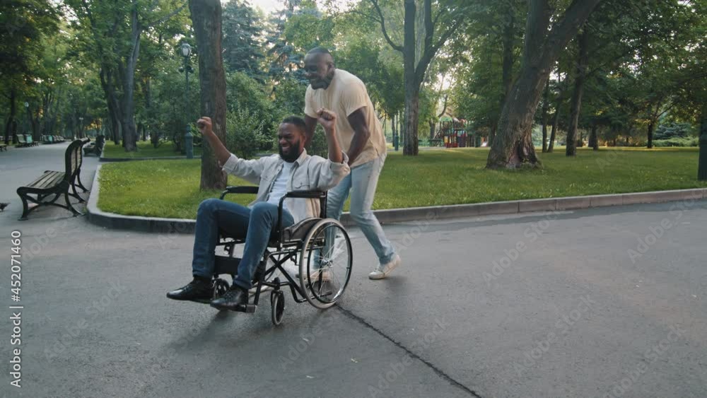 Smiling happy african american young guy in wheelchair having fun with father, raising hands yelling yes recovering from injury received good news. Black man pushing rotates son on chair with wheels Smiling happy african american young guy in wheelchair having fun with father, raising hands yelling yes recovering from injury received good news. Black man pushing rotates son on chair with wheels