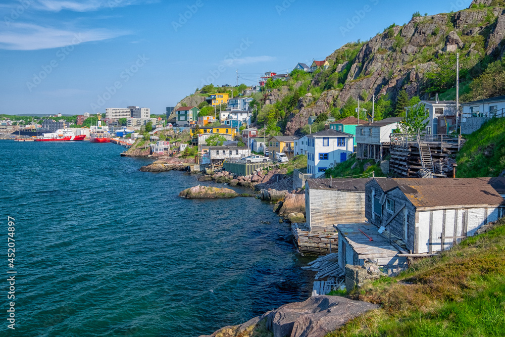 The hillside of St. John's Harbour, Newfoundland, on a sunny day, under blue sky and white clouds. The colorful wooden houses are scattered along the hillside with the blue ocean in the foreground.