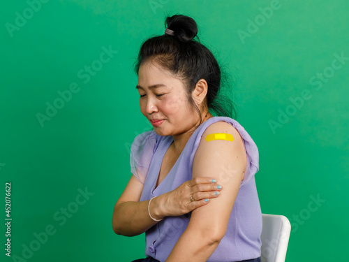 Studio shot of Asian middle aged woman sit smile and show yellow plaster bandage on arm after receive coronavirus covid 19 vaccination by doctor at clinic on green background