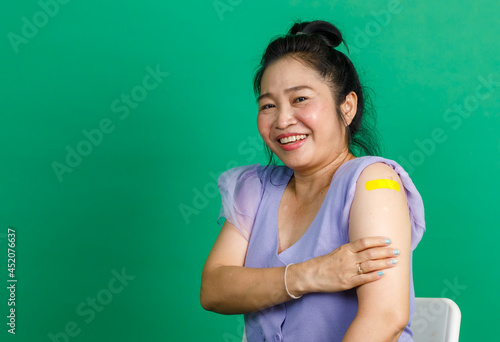 Studio shot Asian happy middle aged bun hair woman patient in purple chiffon dress smiling look at camera showing yellow bandage plaster after receive coronavirus covid 19 vaccine on green background
