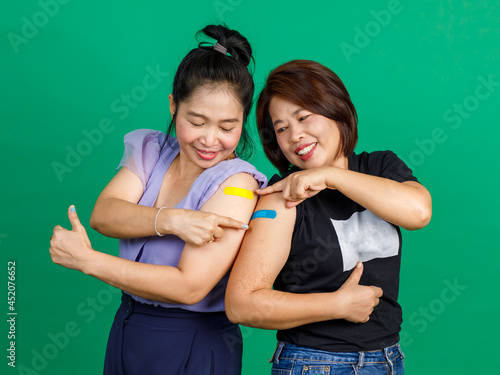 Studio shot of two Asian happy middle aged female patients wear face mask standing look at camera showing thumb up pointing colorful bandage plaster after receiving coronavirus covid 19 vaccine