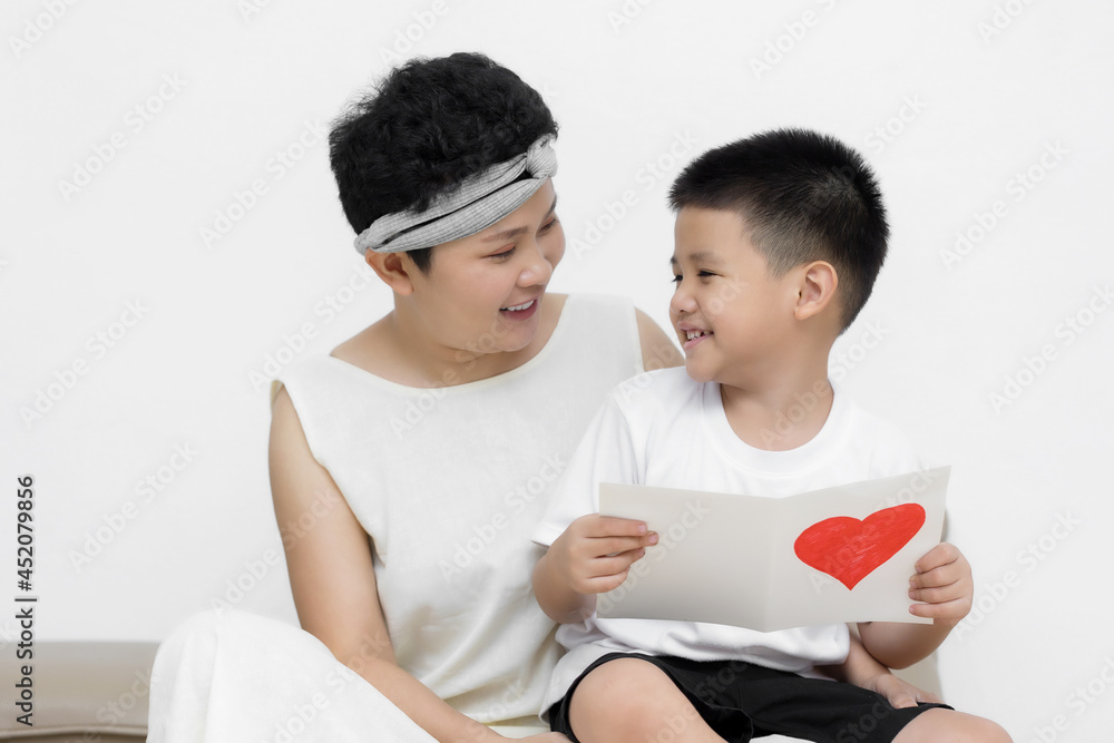 Cute Asian little boy son congratulating his mom happy with Mothers day, giving her handmade greeting postcard with red heart while sitting together on sofa at home. Family holidays concept Cute Asian little boy son congratulating his mom happy with Mothers day, giving her handmade greeting postcard with red heart while sitting together on sofa at home. Family holidays concept