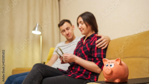 Young family of man and woman sits on yellow sofa and wife counts dollar banknotes, planning budget and giving bill to wife by piggy bank closeup. Woman hiding money in shirt breast-pocket.