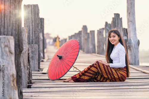 A young Burmese woman wearing traditional clothes with red umbrella at  U Bein teak bridge on Taungthaman lake in Mandalay Division. Myanmar. photo