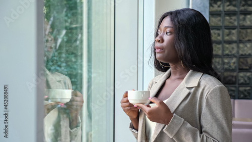 Pretty long haired African-American woman in stylish jacket drinks coffee looking out of large window having break in company office photo