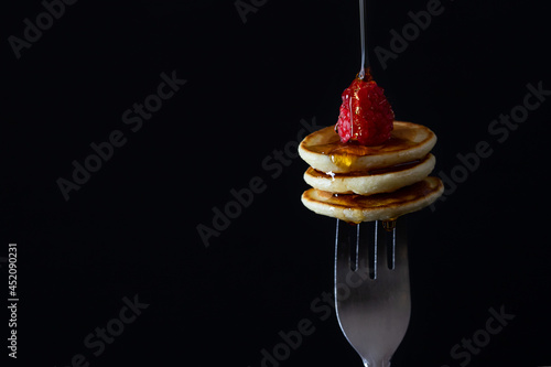 Homemade tiny pancakes with pouring syrup and  raspberry on a fork with a black background, copy space horizontal