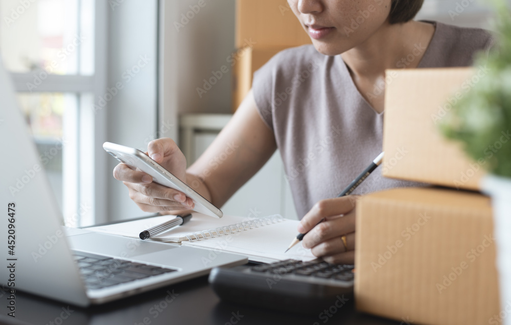 Fototapeta premium Small business, E-commerce, online market concept. Smiling young asian woman entrepreneur checking customer's order on mobile phone and taking note