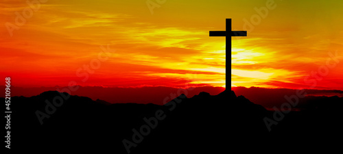 Religious grief landscape background banner panorama - View with black silhouette of mountains, hills, forest and cross / summit cross, in the evening during the sunset, with orange colored sky....