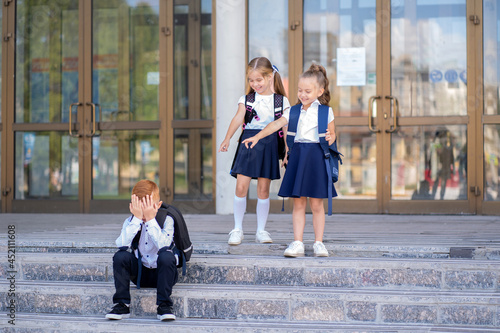 Two little schoolgirls tease a boy with red hair. A student sits on the stairs next to the school and cries while classmates laugh at him.