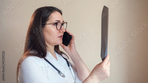 Skilled woman doctor with glasses and stethoscope talks with patient on modern mobile phone looking at X-ray picture on beige background closeup photo
