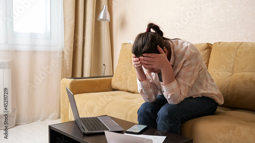 Upset woman tries to calculate high payments on modern mobile phone and holds head in frustration sitting on beige sofa in light living room