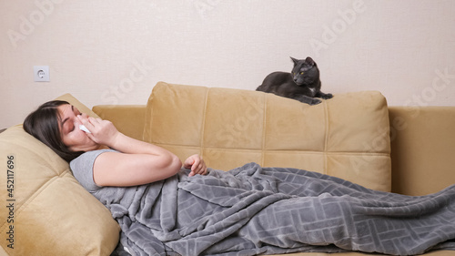 Young woman with long loose hair suffering from allergies sneezes into napkin, lying on sofa under grey blanket near black cat on large pillow at home