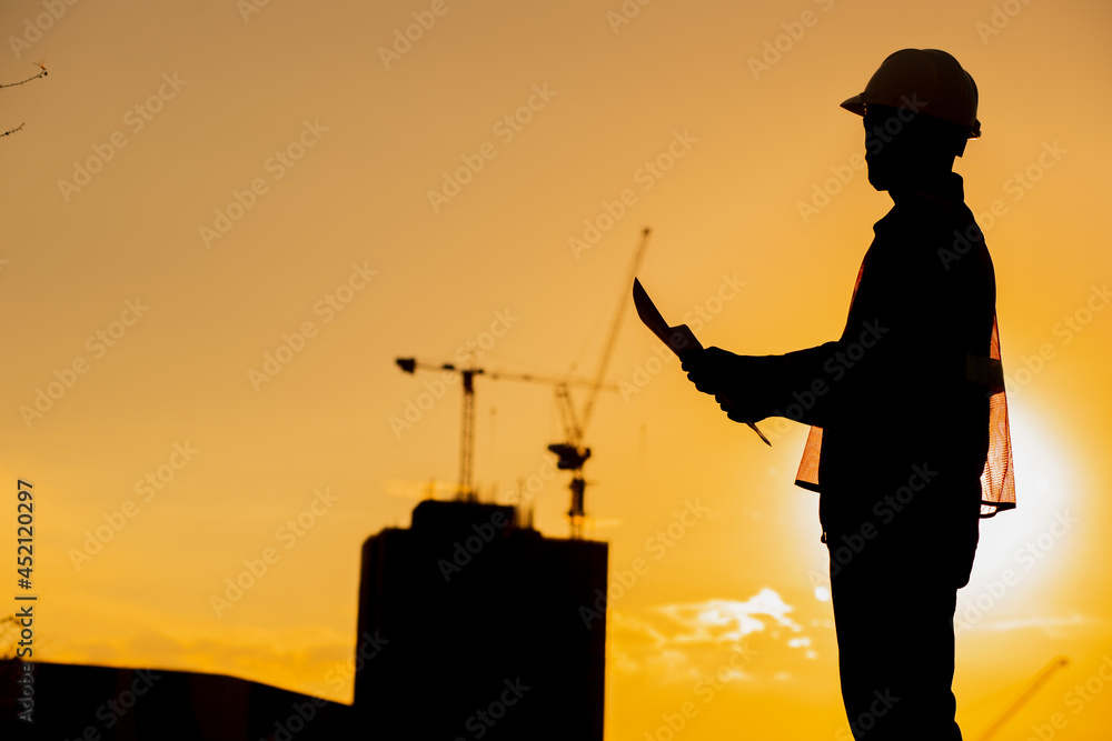 Silhouette of an engineer holding a yellow safety helmet with a tall apartment building and a construction crane on the background of the evening sunset. evening sky
Silhouette of an engineer holding a yellow safety helmet with a tall apartment building and a construction crane on the background of the evening sunset. evening sky