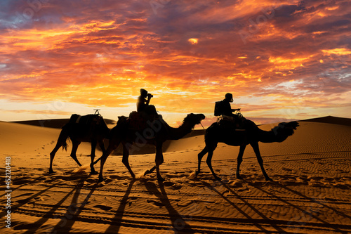 (Selective focus) Stunning sunset behind the silhouette of two tourists riding two camels on the dunes of the Merzouga desert in Morocco. Merzouga is a small village in southeastern Morocco.