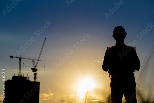 Silhouette of an engineer holding a yellow safety helmet with a tall apartment building and a construction crane on the background of the evening sunset. evening sky 