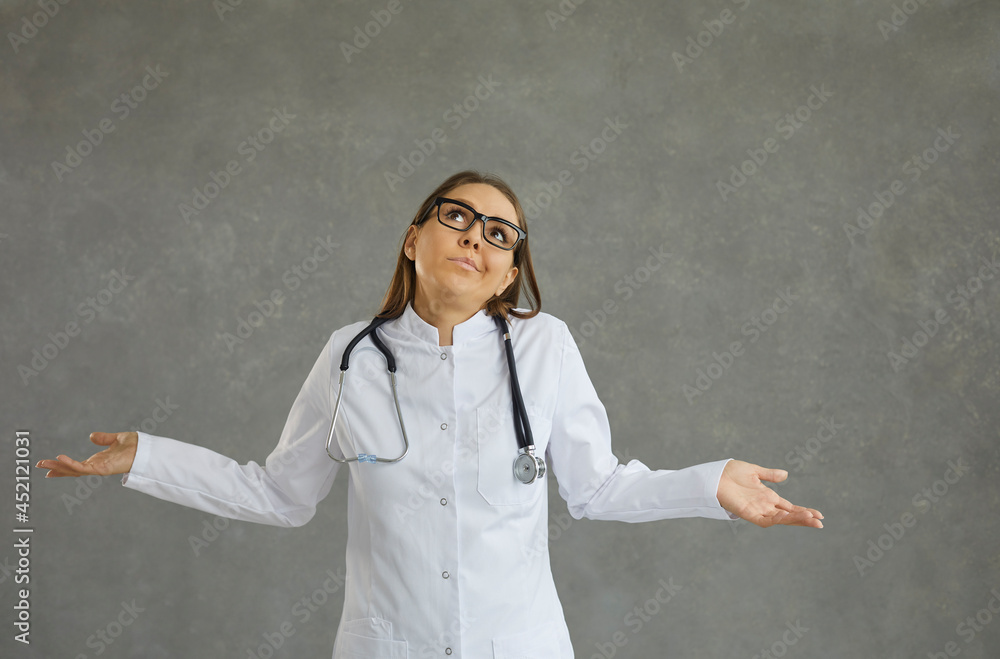 Portrait of a frivolous young female doctor who has no answer and does not know the correct diagnosis. Woman in a medical gown spreads her arms while standing on a gray background. Medicine concept. - obrazy, fototapety, plakaty 