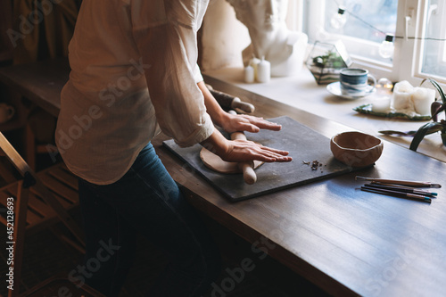 Young attractive woman in white shirt ceramic artist decorating clay plate with tool at table in pottery workshop. Handmade work student, freelance small business, hobbies and crafting