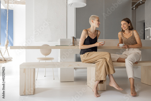 Принти на полотні Lesbian couple drinking coffee, sitting together and talking at modern kitchen in studio apartment