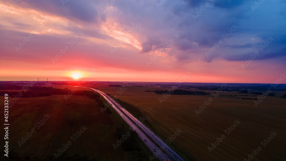 Aerial view of highway on red sunset. Landscape with road near countryside fields. Beautiful winding road leading through rural countryside with evening sunlight. Dramatic sky background. Aerial view of highway on red sunset. Landscape with road near countryside fields. Beautiful winding road leading through rural countryside with evening sunlight. Dramatic sky background.