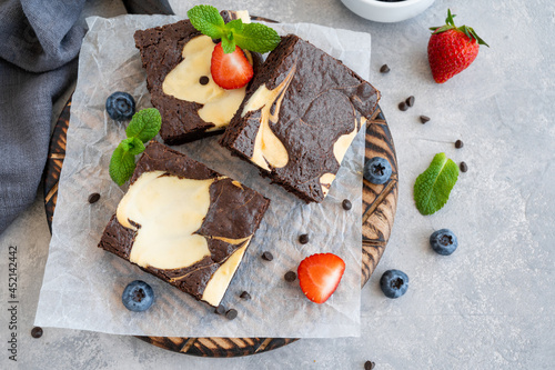 Delicious chocolate brownie cheesecake with fresh berries and mint on a wooden board on a gray concrete background. Copy space.