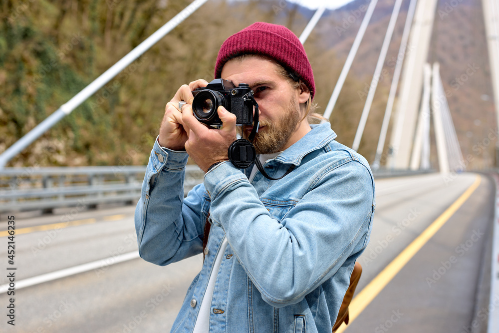 Obraz premium Stylish Caucasian Male Photographer Travel Alone, Walking With Camera. On Bridge. Portrait of handsome guy in denim jacket outdoors, in countryside. copy space.