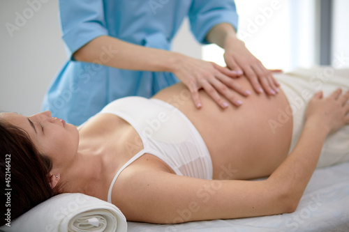 Pregnant young woman with beautiful skin lying on bed having relaxing prenatal massage, physiotherapist doing various techniques for better massaging, focus on brunette mom-to-be