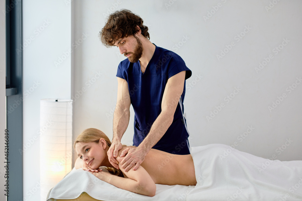 Obraz premium male masseur massaging back and shoulder blades of female lying on massage couch at spa cabinet. Beautiful young blonde woman with perfect skin getting relaxing massage by professional
