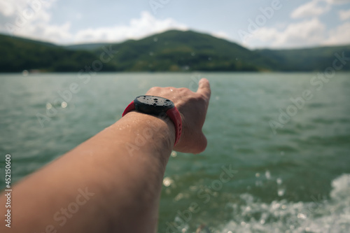 Selective focus on a red running watch with water droplets on the screen and wet man hand with finger pointing towards the mountains across the lake