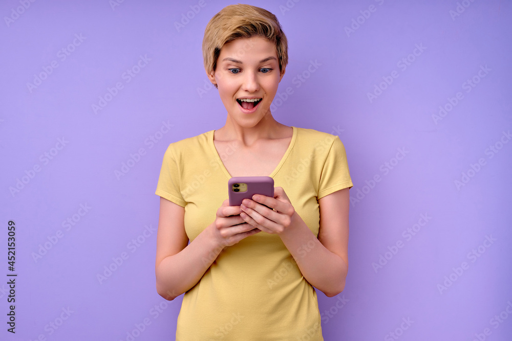 Amazed young woman in casual t-shirt, keeping mouth widely opened,looking at smartphone, caucasian beautiful model posing isolated over purple background, reading shocking news. copy space Amazed young woman in casual t-shirt, keeping mouth widely opened,looking at smartphone, caucasian beautiful model posing isolated over purple background, reading shocking news. copy space