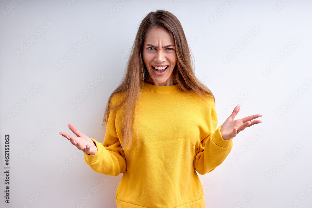 Angry caucasian woman is irritated by something, screaming, spreading arms, looking at camera, isolated on white studio background. Portrait of lady with long hair in yellow sweater. copy space Angry caucasian woman is irritated by something, screaming, spreading arms, looking at camera, isolated on white studio background. Portrait of lady with long hair in yellow sweater. copy space