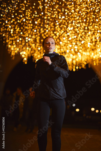 smilm redhead woman in sportswear running along road in city at night illuminated by light garlands lights in background, copy space. portrait of young athlete lady engaged in sport, cardio training