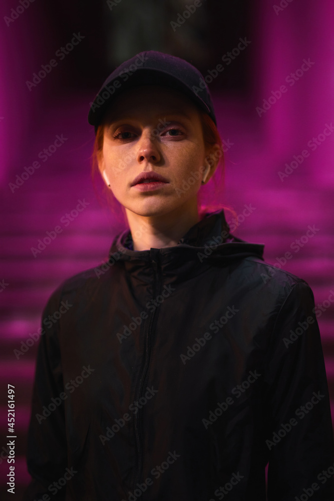 Young caucasian redhead female in black cap, sportive clothes and headphones walking outdoors in night city alone, posing at camera, night life concept. copy space. pink purple light in background