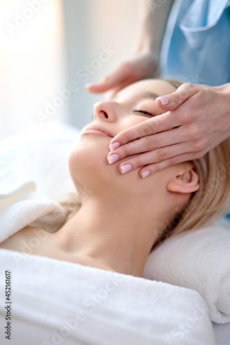 people, beauty, spa, healthy lifestyle and relaxation concept - close up of beautiful young blonde woman lying with closed eyes and having face or head massage in spa salon by professional