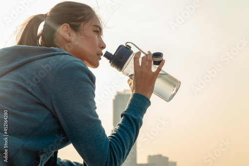 Thirsty young woman runner is resting and drinking water bottle with headphone after morning cardio on street outdoors in the city background.