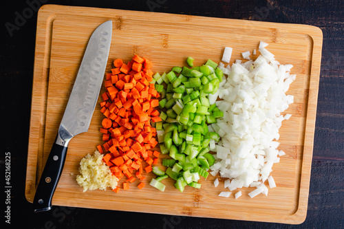 Overhead View of Chopped Vegetables on a Bamboo Cutting Board: Finely chopped carrots, celery, onions, and garlic on a wooden cutting board with a santoku knife