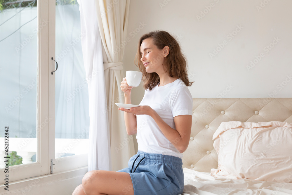 Portrait of beautiful woman drinking morning coffee on bed by the window. Happy beautiful pretty woman relaxing drinking and looking at cup of hot coffee or tea in bedroom. Woman drinking hot coffee. Portrait of beautiful woman drinking morning coffee on bed by the window. Happy beautiful pretty woman relaxing drinking and looking at cup of hot coffee or tea in bedroom. Woman drinking hot coffee.