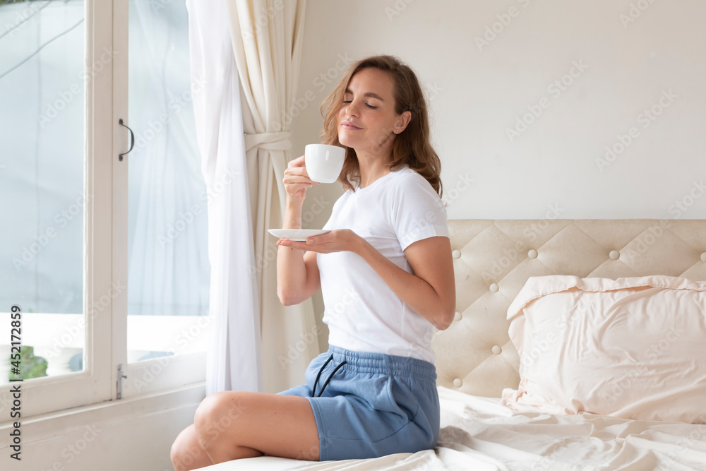 Portrait of beautiful woman drinking morning coffee on bed by the window. Happy beautiful pretty woman relaxing drinking and looking at cup of hot coffee or tea in bedroom. Woman drinking hot coffee. Portrait of beautiful woman drinking morning coffee on bed by the window. Happy beautiful pretty woman relaxing drinking and looking at cup of hot coffee or tea in bedroom. Woman drinking hot coffee.