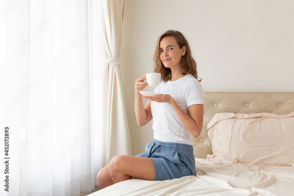Portrait of beautiful woman drinking morning coffee on bed by the window. Happy beautiful pretty woman relaxing drinking and looking at cup of hot coffee or tea in bedroom. Woman drinking hot coffee. Portrait of beautiful woman drinking morning coffee on bed by the window. Happy beautiful pretty woman relaxing drinking and looking at cup of hot coffee or tea in bedroom. Woman drinking hot coffee.