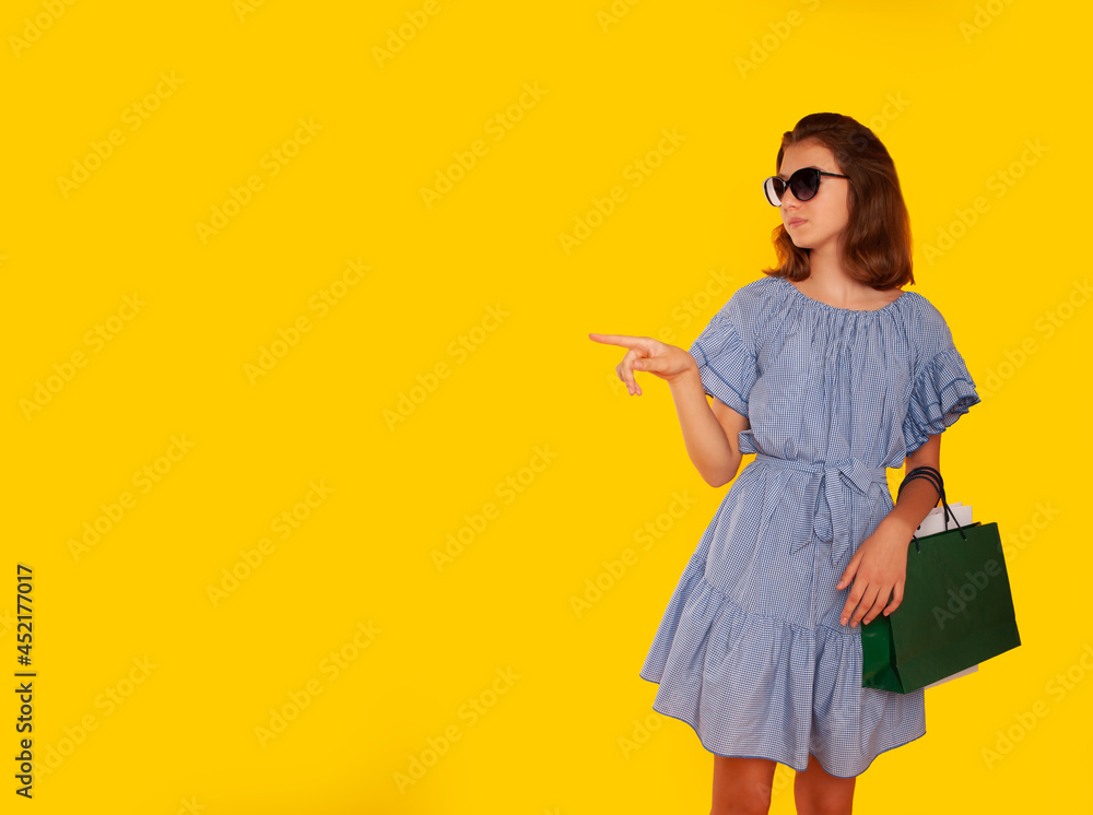girl on a yellow isolated background with green and white packages in her hands in a blue dress and glasses shopping in a mall in a good mood and with an adorable smile. Place for your text. Advertisi girl on a yellow isolated background with green and white packages in her hands in a blue dress and glasses shopping in a mall in a good mood and with an adorable smile. Place for your text. Advertisi