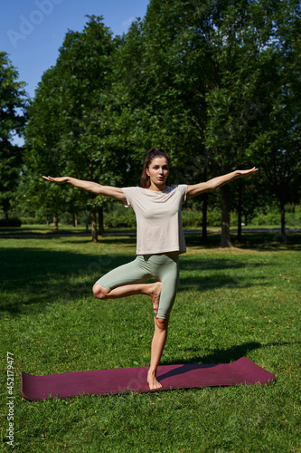 beautiful young woman stands in the park on a sunny day in summer. stands in the pose of a tree (Vrikshasana) on one leg and with arms outstretched. yoga meditation. sports, unity with nature, 