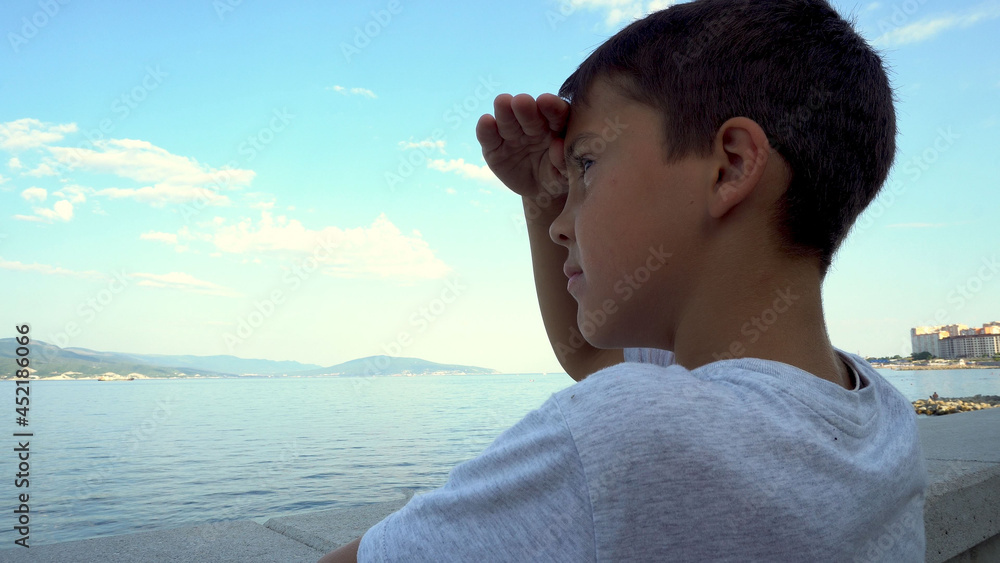 The boy stands on the embankment and looks at the sea. Tanned skin. Age 7-8 years old. Dreamer. Gray T-shirt. Blue sky.