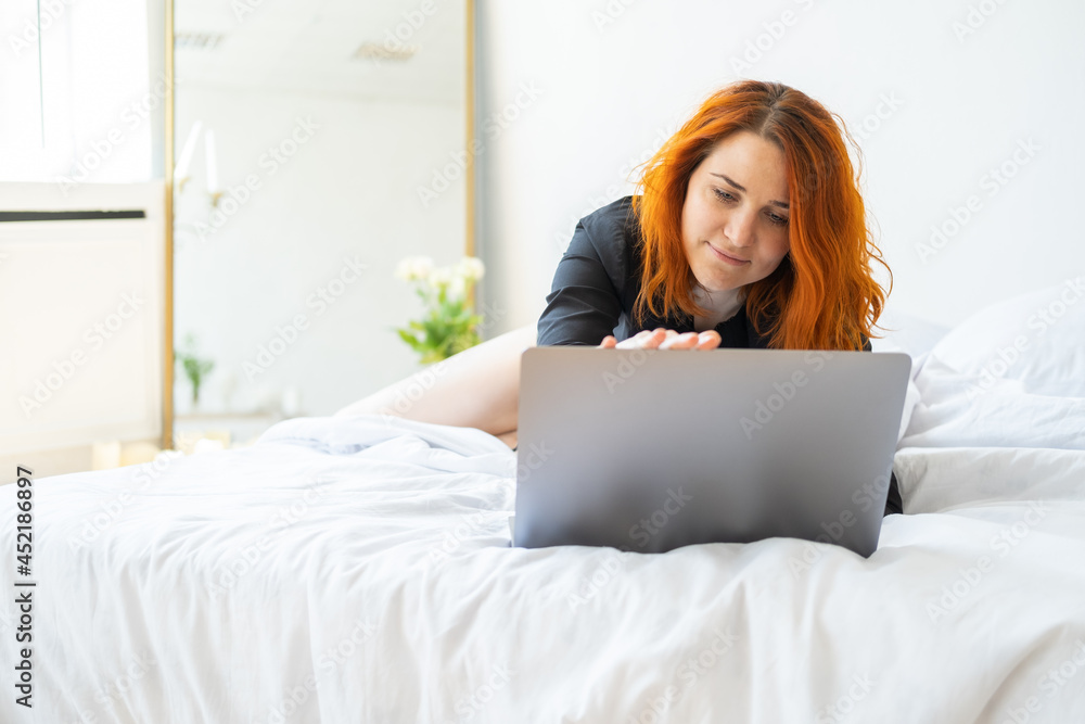 attractive smiling ginger woman middle age lying in bed using laptop in bright room attractive smiling ginger woman middle age lying in bed using laptop in bright room